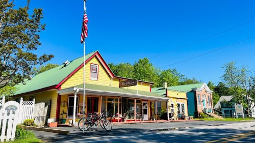 A colorful, quaint storefront with a flag, surrounded by greenery and a clear blue sky. Bicycles are parked outside.