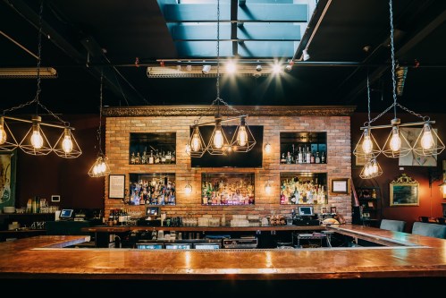 A stylish bar with a wooden counter, brick wall, and illuminated shelves filled with various bottles.