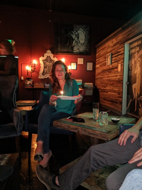 A woman sits at a table in a dimly lit bar, holding a plate with a lit candle, surrounded by drinks and decor.
