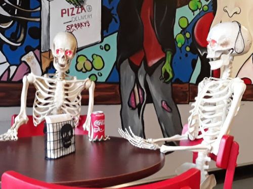 Two skeletons sitting at a table with a soda can, surrounded by colorful, spooky wall art.