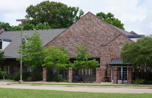 A brick building with a sloped roof, surrounded by trees and greenery, under a partly cloudy sky.