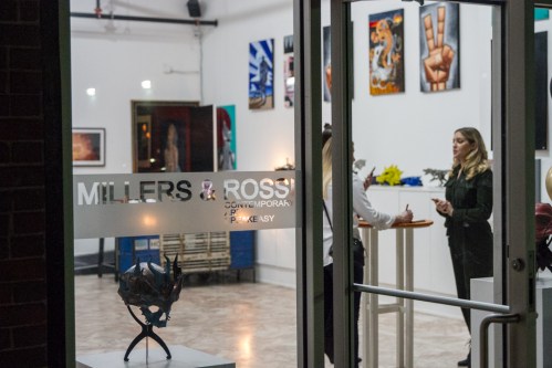 Two people converse inside an art gallery, with various artworks displayed on the walls.