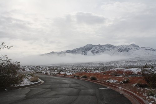 A winding road leads through a snowy landscape with mountains and low-hanging clouds in the background.