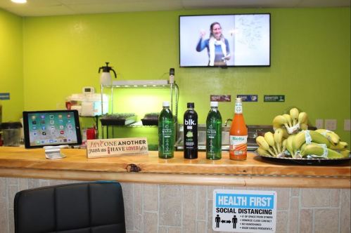 A counter with drinks, bananas, and a tablet, featuring a woman presenting in the background. Health guidelines are displayed.