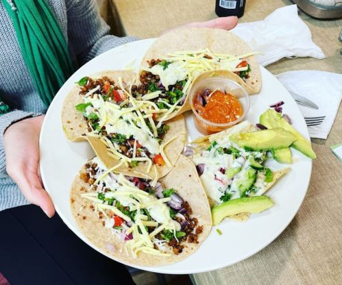 A plate of tacos filled with meat, vegetables, and toppings, served with a side of sauce and avocado slices.