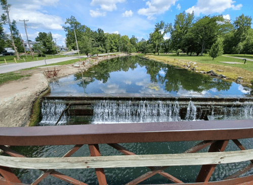 A serene view of a small waterfall over a dam, reflecting trees and blue sky in calm water.