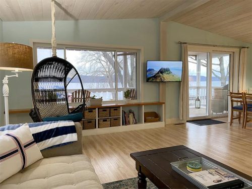 Cozy living room with a hanging chair, TV, and large windows overlooking a lake. Light wood flooring and soft decor.