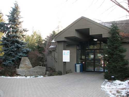 Entrance of a nature center with a stone sign, surrounded by trees and a snow-dusted pathway.