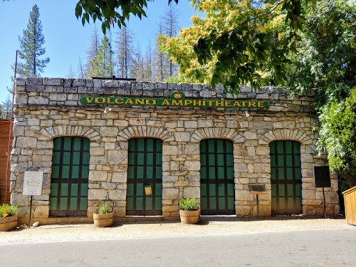 Stone building with green doors and "Volcano Amphitheatre" sign, surrounded by trees and plants.