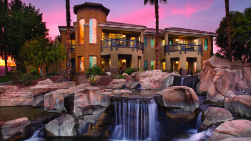 A luxurious two-story home with a waterfall and palm trees, set against a vibrant sunset sky.