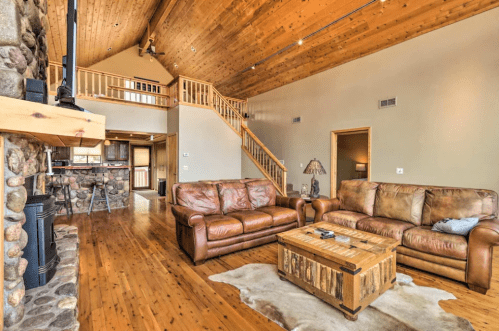 Cozy living room with leather sofas, wooden accents, and a stone fireplace, featuring a staircase and rustic decor.