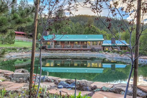 A rustic log cabin with a green roof reflects in a serene pond, surrounded by trees and mountains.