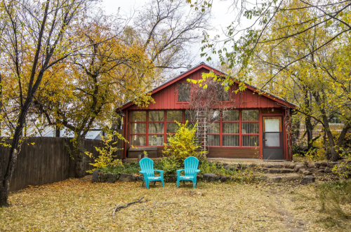 A cozy red cabin surrounded by autumn trees, with two blue chairs in the foreground on a leaf-covered yard.