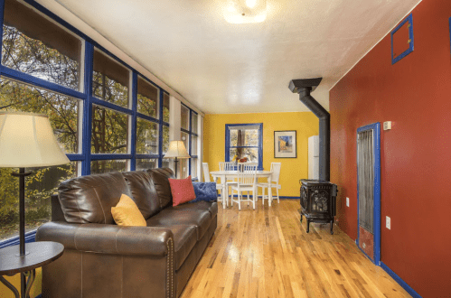 Cozy living space with large windows, a brown leather sofa, dining table, and colorful walls in warm tones.