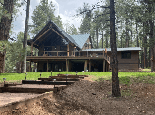 A modern cabin nestled among tall pine trees, featuring a large porch and a sloped roof. Pathway leads to the entrance.