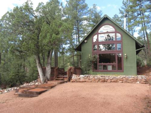 A green cabin with large windows surrounded by trees and a gravel path, featuring a wooden deck and stone steps.