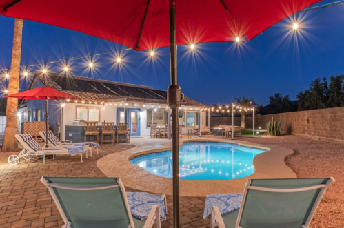 A serene backyard at night featuring a pool, lounge chairs, and string lights above a cozy home.