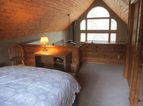 Cozy attic bedroom with a bed, wooden furniture, a lamp, and large windows letting in natural light.