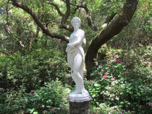 A white marble statue of a woman stands gracefully among lush greenery and blooming flowers.