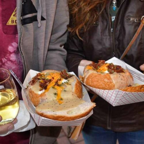 Two people hold bowls of bread filled with creamy soup and toppings, with a glass of drink nearby.
