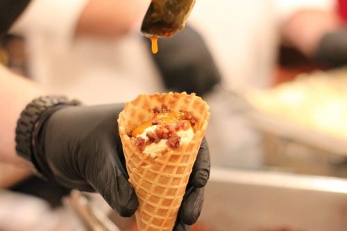 A hand in a black glove holds a waffle cone filled with a savory mixture, with sauce being drizzled on top.