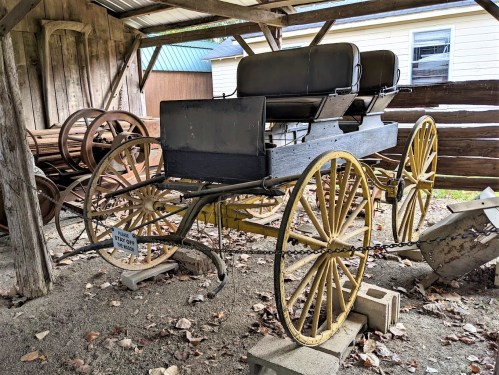 An old horse-drawn carriage with yellow wheels, resting under a wooden shelter surrounded by rustic artifacts.
