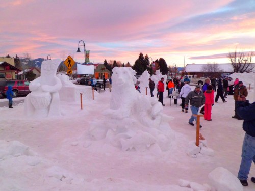A snowy scene with people admiring large snow sculptures against a colorful sunset sky.