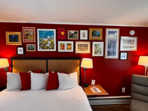 A cozy hotel room with a red wall, a bed with white and red pillows, and a gallery of framed artwork above the headboard.