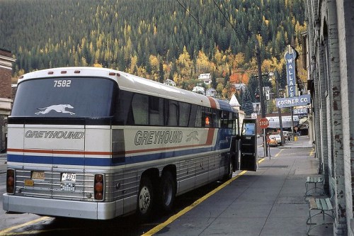 A Greyhound bus parked on a street with autumn trees in the background and vintage storefronts nearby.
