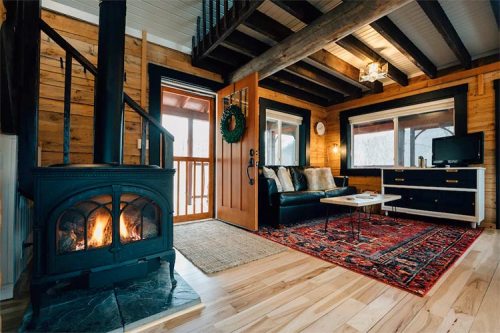 Cozy cabin living room with a wood stove, plush sofa, coffee table, and a vibrant rug, featuring wooden beams and large windows.