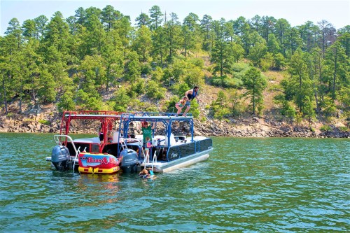 A group of people on a boat at a lake, with one person jumping off and another swimming nearby, surrounded by trees.