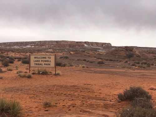 A sign reading "Welcome to Lake Powell Tribal Park" in a desert landscape with rocky hills and sparse vegetation.