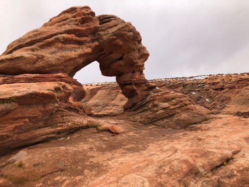 A natural rock formation with a prominent arch, surrounded by rugged terrain and a cloudy sky.