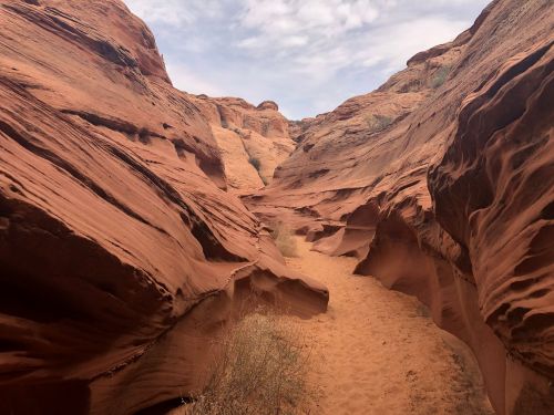 A narrow canyon with smooth, reddish rock formations and a sandy path, under a cloudy sky.
