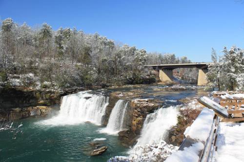 A snowy landscape featuring a waterfall and a bridge under a clear blue sky. Trees are dusted with snow.