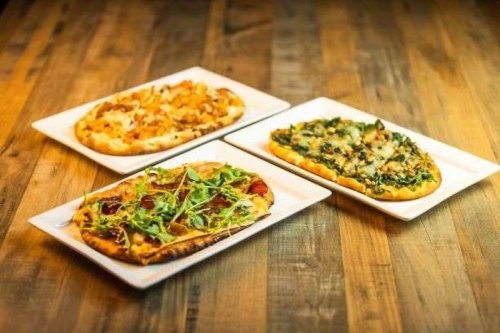 Three flatbreads on white plates: one with chicken, one with greens, and one with spinach and cheese, on a wooden table.
