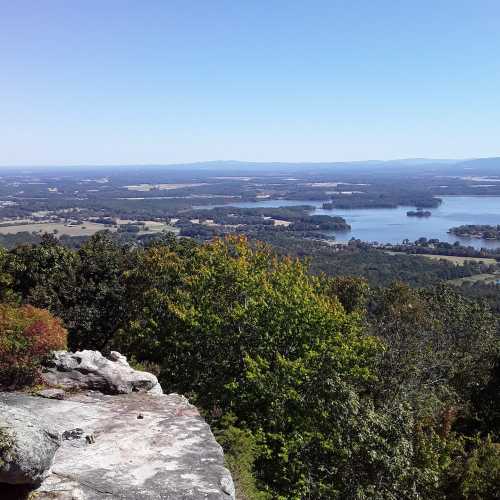 A scenic view from a rocky overlook, showcasing lush green hills and a calm body of water under a clear blue sky.