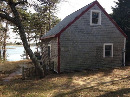 A small, shingled cottage with a red trim, surrounded by trees and overlooking a calm body of water.