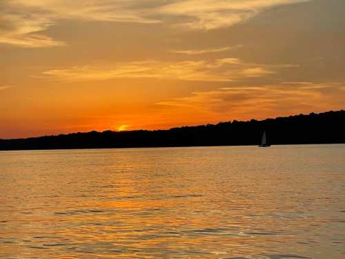 A serene sunset over a calm lake, with a silhouette of a sailboat and trees along the horizon.