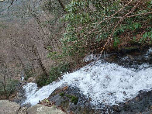 A cascading waterfall flows over rocks, surrounded by bare trees and greenery in a serene natural setting.