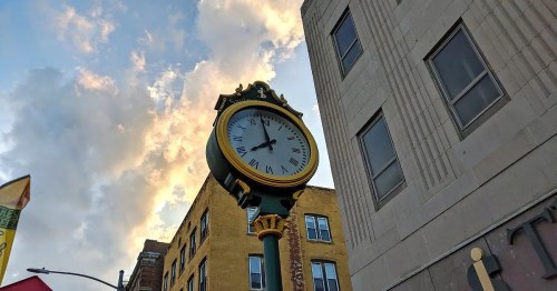 A vintage clock tower against a cloudy sky, with buildings on either side.