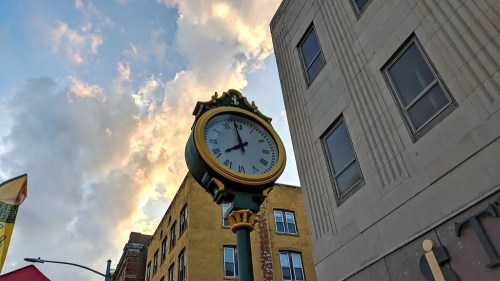 A large clock mounted on a building, with a colorful sky and clouds in the background.