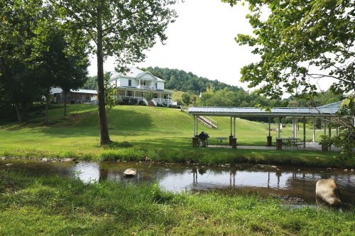 A scenic view of a house on a hill, surrounded by trees and a stream in the foreground.