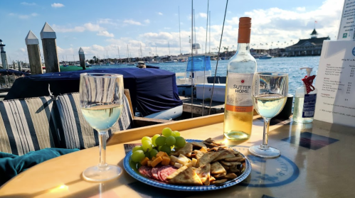A scenic waterfront table with two glasses of white wine, a plate of snacks, and boats in the background under a blue sky.