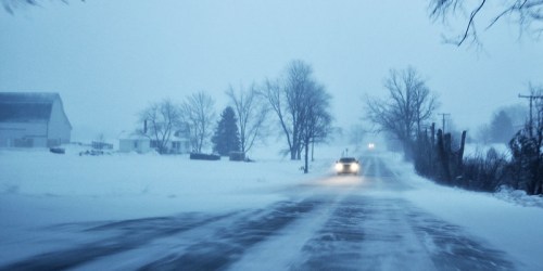 A snowy road with a car driving through a winter storm, surrounded by trees and distant buildings.