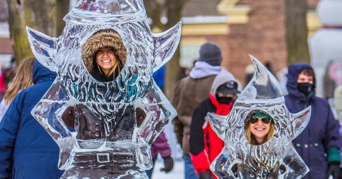 Two women stand inside large ice sculptures shaped like whimsical characters, smiling amidst a winter festival crowd.
