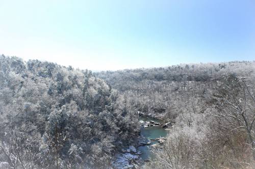 A snowy landscape with trees covered in frost, overlooking a river winding through a serene valley under a clear blue sky.