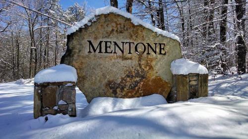 A large stone sign reading "MENTONE" covered in snow, surrounded by trees in a winter landscape.