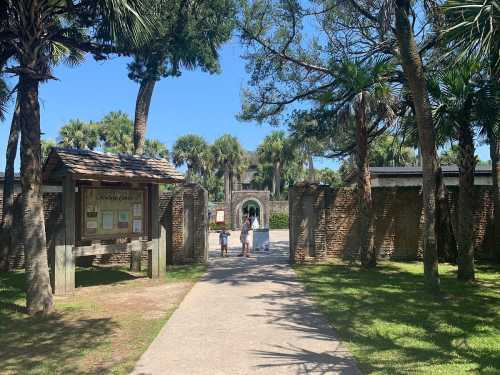 A pathway leads through palm trees to a historic site entrance, with visitors walking towards it.