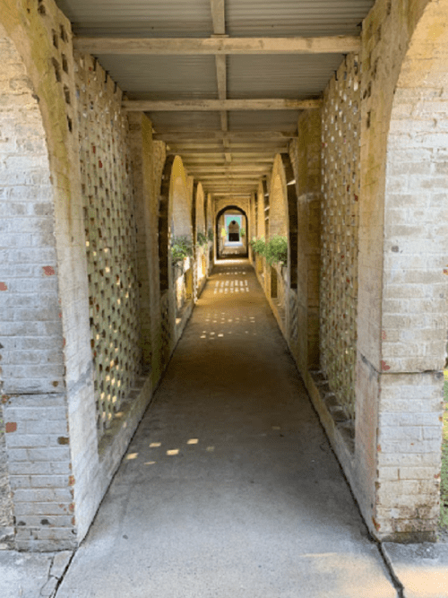 A long, arched corridor with brick walls and latticework, leading to a bright opening at the end.